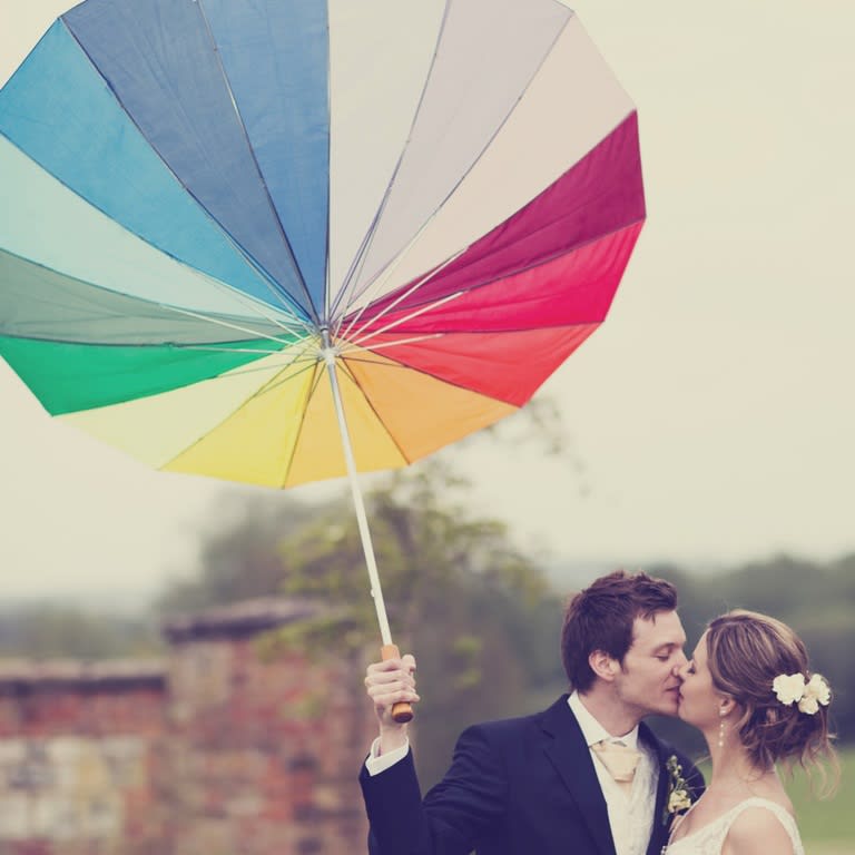 Bridebook.co.uk- bride and groom kissing under multicoloured umbrella