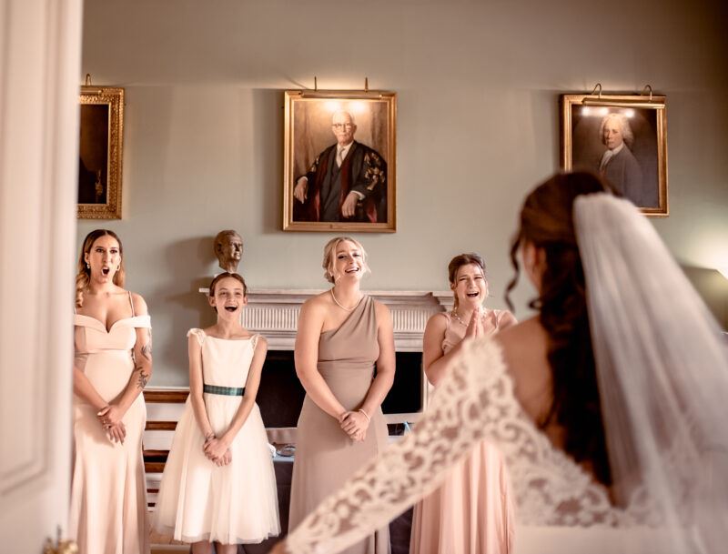 Four bridesmaids stand in a row in the room of a stately home, each reacting differently to seeing her in her wedding dress for the first time. The bride stands with her back to the camera and her arms spread out and her veil visible.