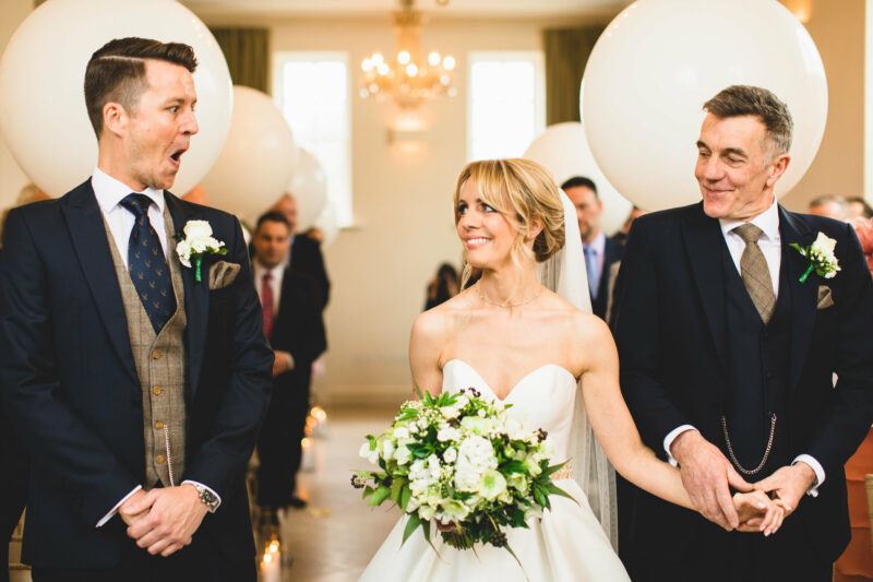 A groom stands on the left, his mouth open in shock at seeing his bride standing next to him in her wedding dress, with the bride's father holding her hand on the right. Behind them in the aisle are white balloons and a golden chandelier.