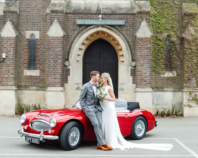 Bridebook.co.uk- bride and groom smiling next to red wedding car