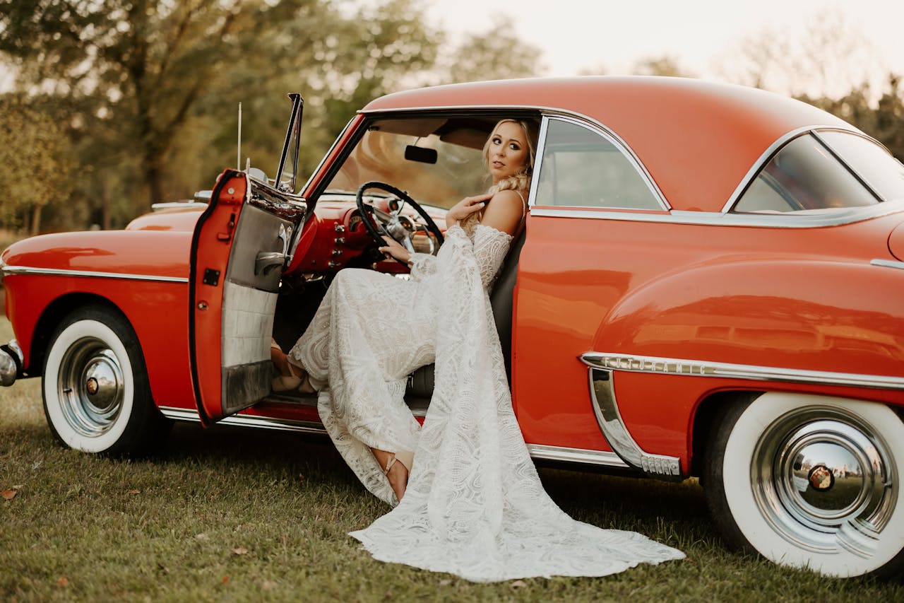 Une mariée en robe blanche bohème assise dans une voiture rouge vintage, dégageant une élégance rétro et une ambiance romantique.