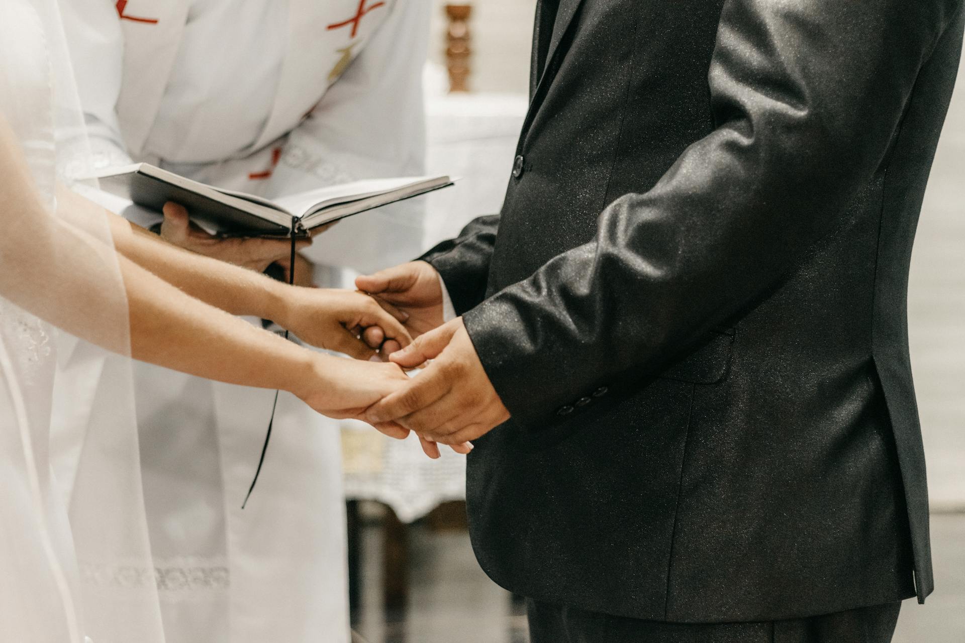 Gros plan sur les mains d’un couple lors d’une cérémonie religieuse, avec un prêtre en tenue blanche tenant un livre ouvert