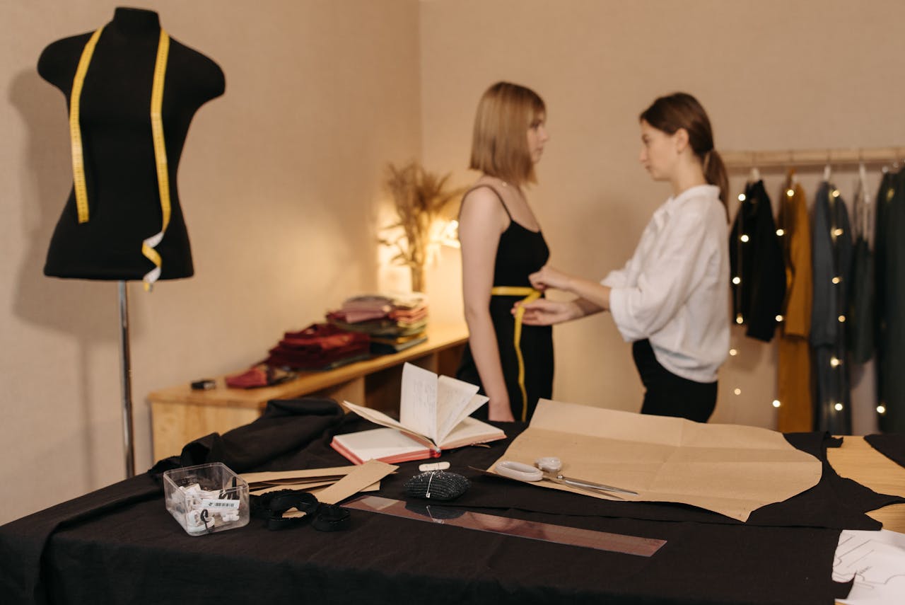 Une couturière mesure le tour de taille d’une cliente dans un atelier de couture, avec un mètre ruban jaune, un mannequin et du matériel de couture en arrière-plan.
