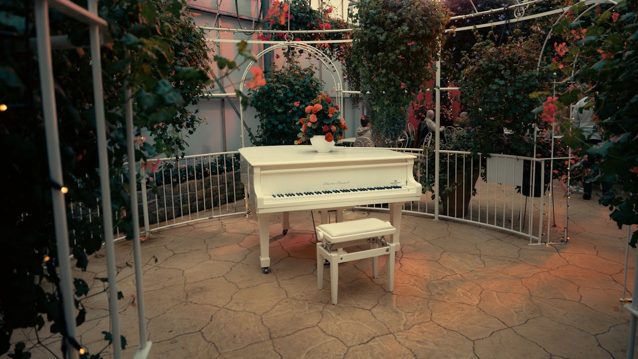 Un piano à queue blanc décoré de fleurs rouges, placé sous une pergola entourée de verdure et illuminée de petites lumières.