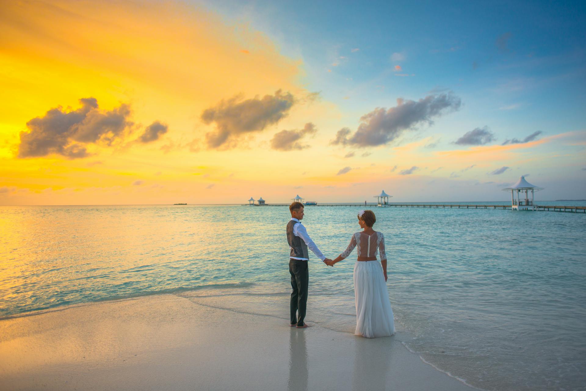 Un couple en tenue de mariage se tenant la main sur une plage au coucher du soleil, face à l’océan et à un ponton avec des pavillons blancs.