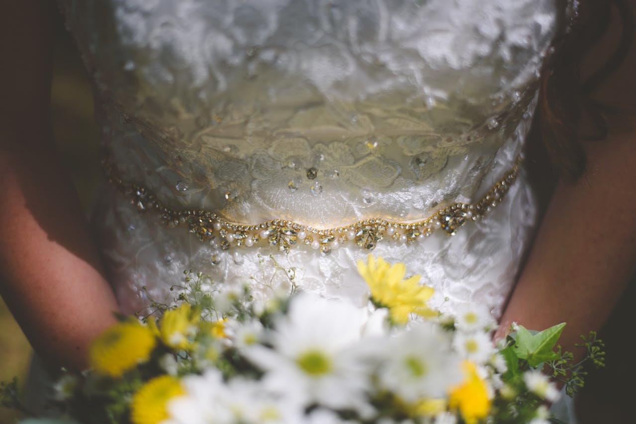  Gros plan sur une robe de mariée en dentelle avec une ceinture perlée dorée et un bouquet de fleurs blanches et jaunes en premier plan.