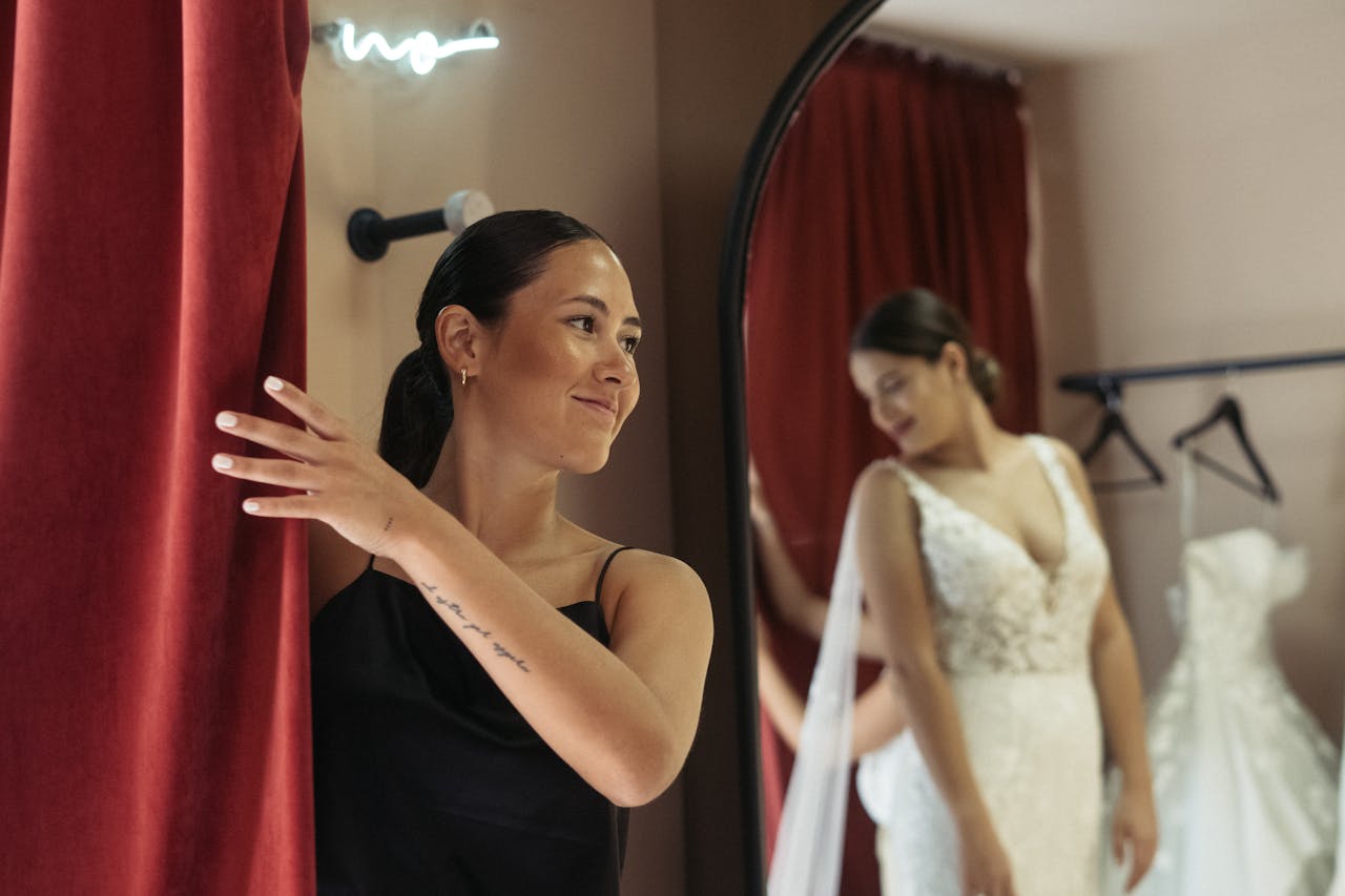 Une future mariée en robe blanche élégante se regarde dans un miroir lors d'un essayage, tandis qu'une amie souriante ouvre le rideau de la cabine d'essayage.