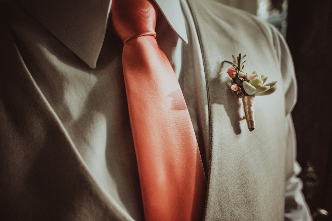 Détail d’un costume beige avec une cravate corail et une boutonnière florale, illustrant un style naturel et raffiné pour un mariage champêtre.