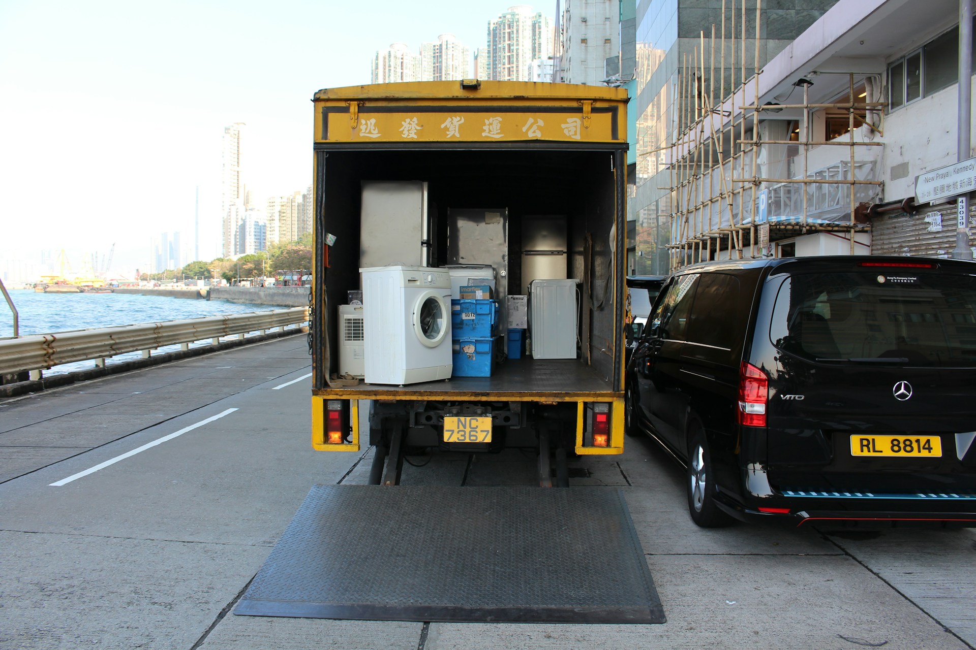 Camion de déménagement jaune avec sa porte arrière ouverte, chargé d'appareils électroménagers et de boîtes, stationné près d'un van noir sur une route en bord de mer avec des immeubles en arrière-plan.