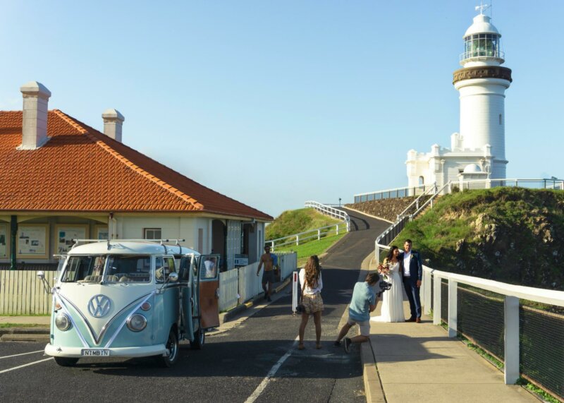 Couple de mariés en train de faire des photos devant un phare, à côté d'un van Volkswagen